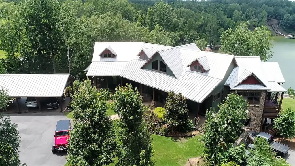 Aerial view of lake house surrounded by tall trees with a light metal ceiling and cars parked in the driveway